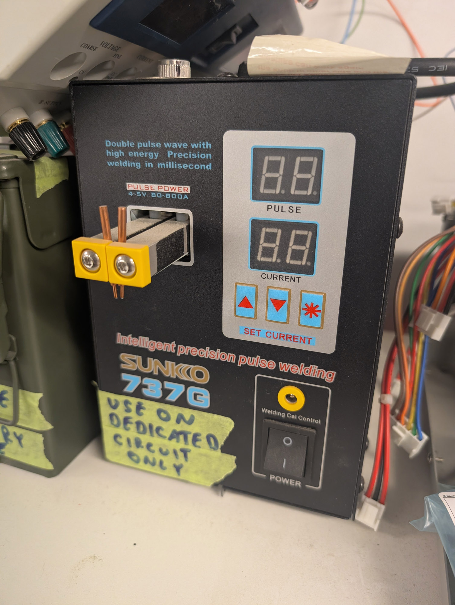A black metal box with a control panel on the front. The front has 2-digit 7-segment displays for Pulse and Current, with buttons for up, down, and an asterisk below that. The left side has two large busbars coming out of the front, with pointed tips attached to them. Above that it says "Pulse Power - 4-5V, 80-800A". There is a black rocker power switch and a terminal labelled "Welding Cal Control" at the bottom, and masking tape that says "Use on dedicated circuit only"